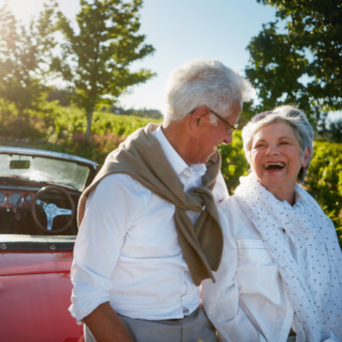 An older couple smiles and laughs together beside a red convertible on a sunny day, surrounded by trees and greenery.