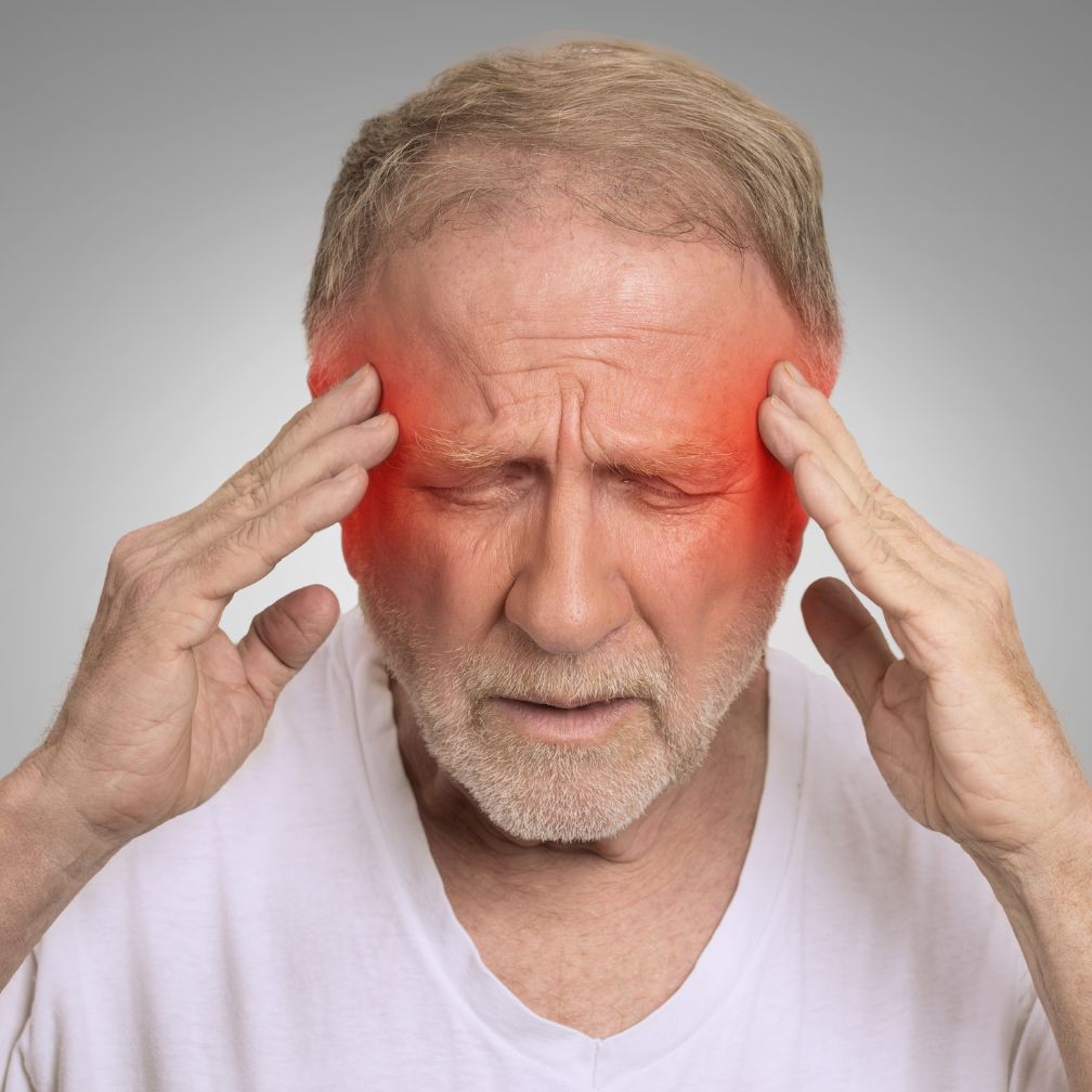Older man with closed eyes holds his temples, grimacing in pain, with red highlights on both sides of his head, indicating a headache or migraine. He is wearing a white shirt and the background is plain gray.