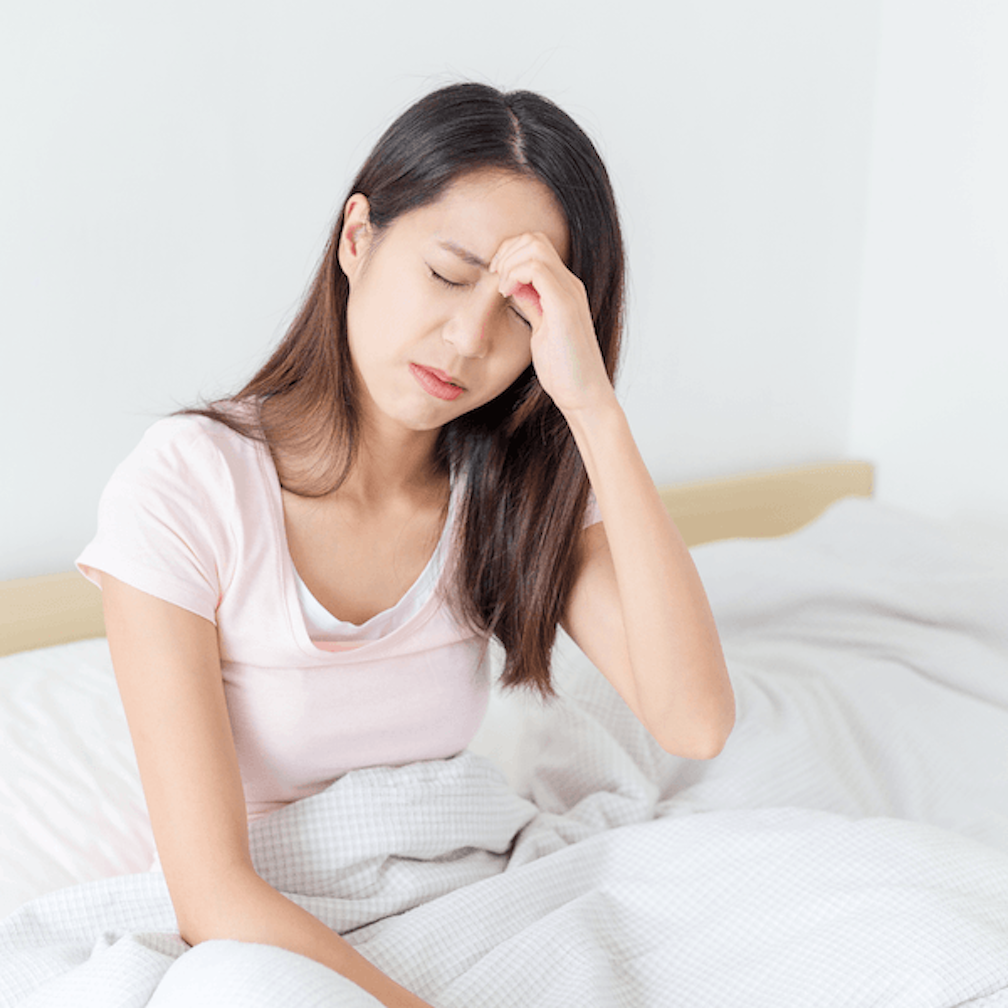 A woman sits on a bed with her eyes closed, holding her forehead as if she has a headache or feels unwell. She is wearing a light pink shirt and is partially covered by a white blanket.