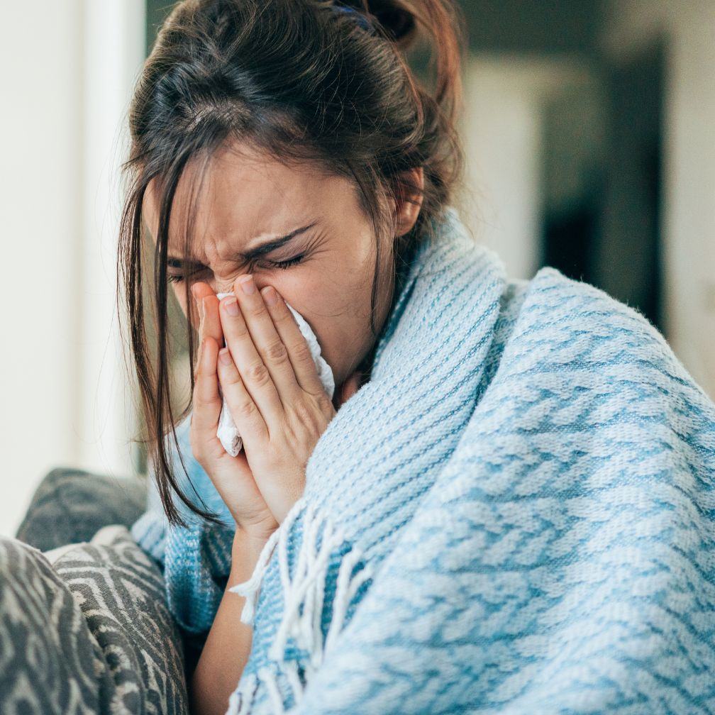 A woman wrapped in a blue blanket sits on a couch, holding a tissue to her nose and appearing to sneeze or blow her nose.