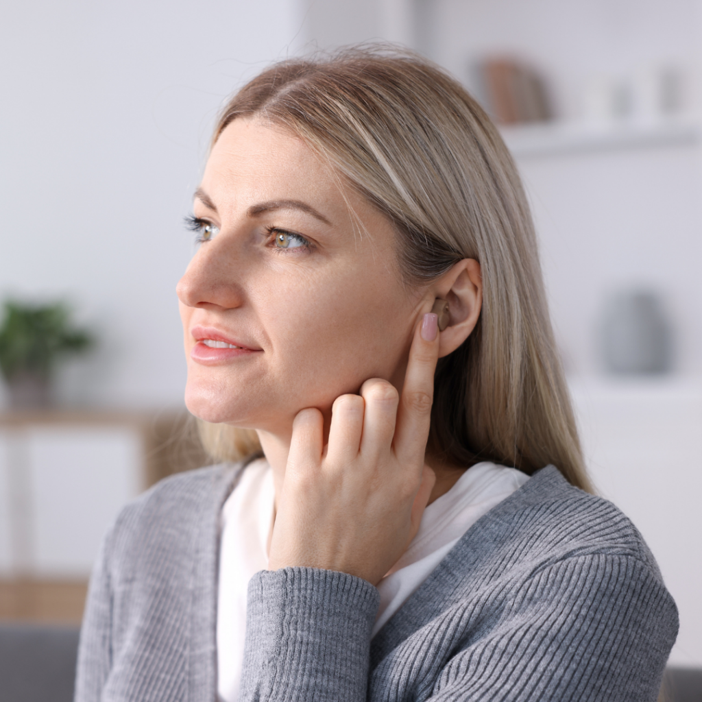 A woman with blonde hair points to her ear with her finger while looking thoughtfully to the side, wearing a gray sweater in a softly lit room.