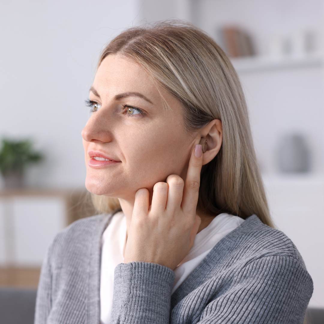 A woman with blonde hair points to her ear with her finger while looking thoughtfully to the side, wearing a gray sweater in a softly lit room.