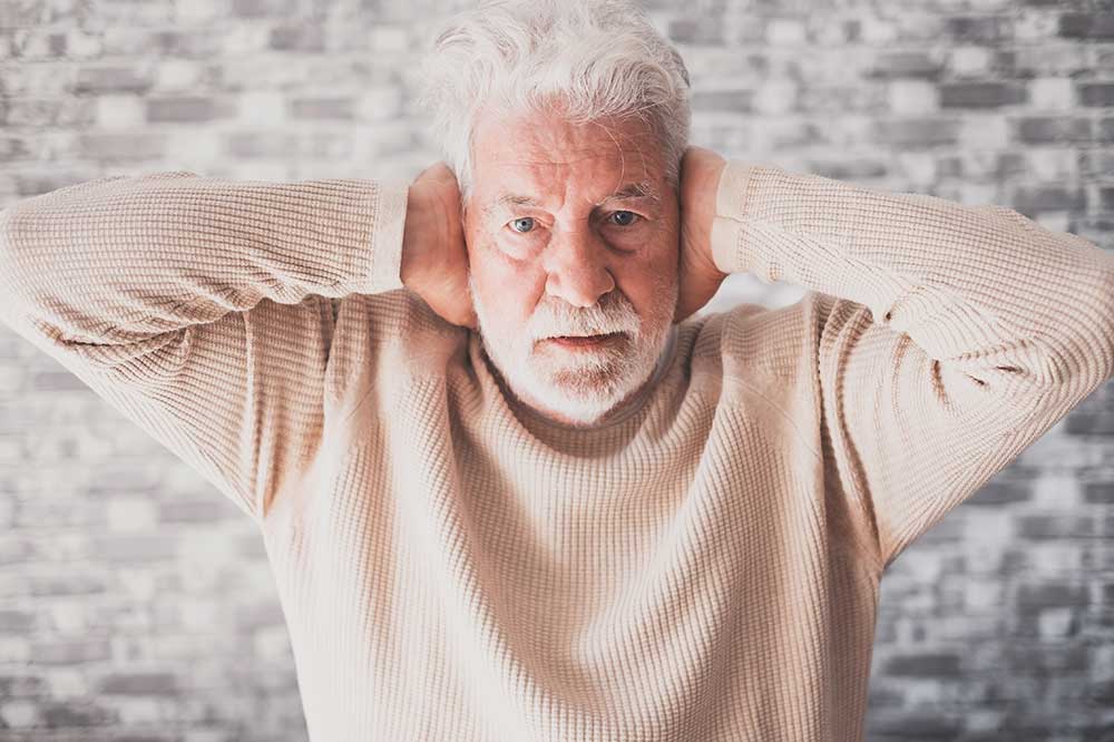 Older man with a white beard covering both ears over a clogged ear feeling, a concern at Enhanced Hearing Specialists in Dickson City, PA